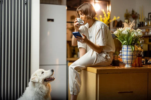 年轻女人在家里和她的狗坐在厨房里 一边用智能手机一边喝咖啡 上午例行公事和家庭生活方式的概念 — 图库照片