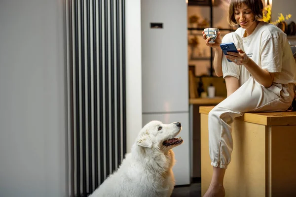 年轻女人在家里和她的狗坐在厨房里 一边用智能手机一边喝咖啡 上午例行公事和家庭生活方式的概念 — 图库照片