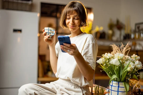 年轻女人用智能手机喝咖啡 早上在漂亮的厨房里呆着 家里开着鲜花 — 图库照片