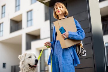 Genç bir kadın, köpeğiyle birlikte apartmanın yakınında duran otomatik postane makinesinden paketleri alıyor. Hızlı taşıma ve kentsel yaşam tarzı kavramı