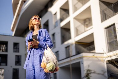 Genç bir kadın, alışveriş torbalarıyla dolu bir çantayla eve yürürken telefon kullanıyor. Sürdürülebilir ve kentsel yaşam tarzı, alışveriş gıda konseptinde yeniden kullanılabilir çanta kullanımı