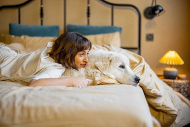 Cute young woman hugs with her white adorable dog while lying on bed at home. Concept of friendship with pets and home coziness