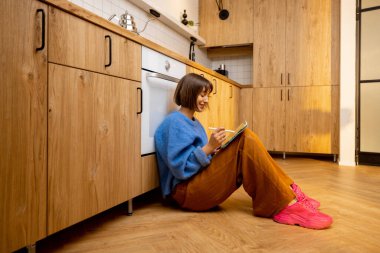 Genç bir kadın, yeni ve şık bir stüdyo dairede mutfak zemininde dijital tabletle oturuyor. Uzaktan çalışma ya da evde boş zaman kavramı