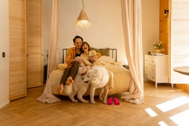 Genç çift yatakta birlikte oturup köpekle oynayarak eğleniyor. Boş zamanlarını evlerinin rahat yatak odasında mutlu geçiriyorlar.