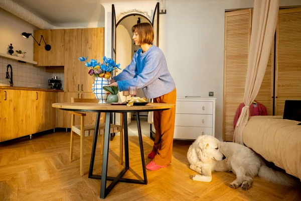 女人用花瓶装饰餐桌 在现代风格的工作室公寓里做家务活 狗躺在附近 — 图库照片