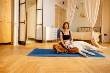 Genç bir kadın sevimli köpeğini önemsiyor, evde yoga yaparken birbirine sarılıyor. Köpek terapisi ve akıl sağlığı kavramı