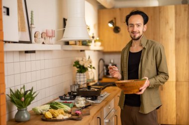 Yakışıklı adam, şık ev mutfağında sağlıklı vejetaryen yemekleri pişirirken ahşap kasede salata karıştırıyor.