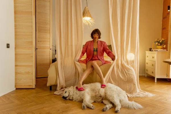 穿着粉红西服的时尚女人坐在舒适的床上 头戴天篷 脚紧抱着睡狗 用米色的语调坐在舒适的卧室里 — 图库照片