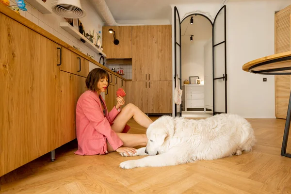 身穿粉色西服的女人在厨房里和她的大白狗一起吃着羊角面包 家庭奢侈生活和与宠物的友谊概念 — 图库照片