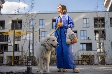 Mavi pijamalı genç bir kadın köpeğiyle yürüyor ve apartmanın iç bahçesinde içi taze meyve ve sebze dolu bir ağ çantası taşıyor. Sürdürülebilir ve modern yaşam tarzı kavramı