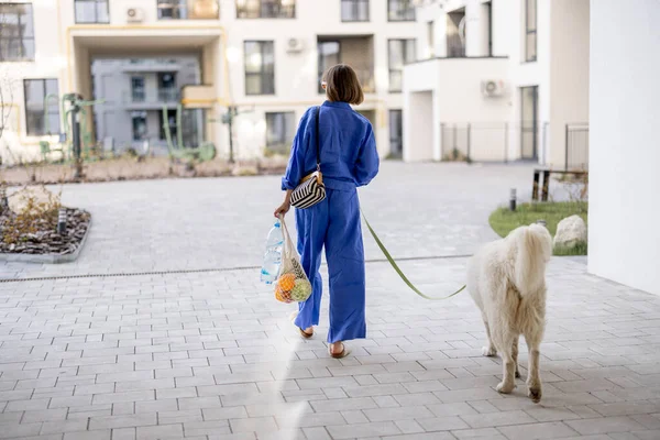 Young woman in blue pajamas walks with her dog and carry mesh bag full of fresh fruits and vegetables at inner yard of apartment building. Sustainable and modern lifestyle concept