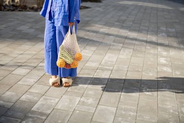 Terlik giymiş bir kadın eve yürürken içi taze meyve ve sebzelerle dolu bir çanta taşıyor, yakın plan. Sürdürülebilir yaşam tarzı ve alışveriş gıda konseptinde yeniden kullanılabilir çanta kullanımı