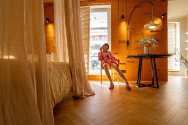 Pembe elbiseli kadın şık ve rahat bir apartman dairesinde yemek masasında rahatça oturuyor.