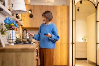 Genç kadın, modern mutfağın içinde telefonla beklerken ocağın üzerindeki çaydanlığı ısıtıyor. Ev eğlencesi ve şık iç mekan kavramı