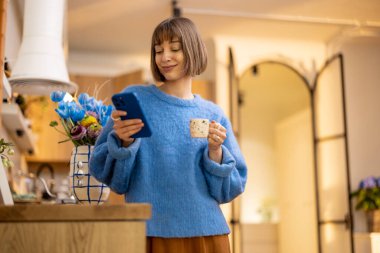 Genç bir kadın evde mutfaktaki akıllı telefona bakarken kahve içmekten hoşlanır. Boş zaman ve ev hayatı kavramı