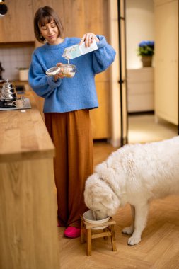 Genç bir kadın, tatlı köpeğiyle mutfaktayken yoğurt ve granola ile kahvaltı hazırlıyor. Evcil hayvan konseptiyle aile hayatı ve arkadaşlık.