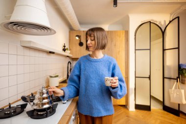 Genç bir kadın, modern mutfağın içinde çaydanlık ve fincanla duran bir kahve içeceği hazırlıyor. Ev hayatı kavramı