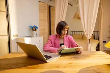 Pembe takım elbiseli genç iş kadını rahat bir ofiste dijital tablet üzerinde çalışıyor. Konforlu ev atmosferinden bağımsız çalışma kavramı