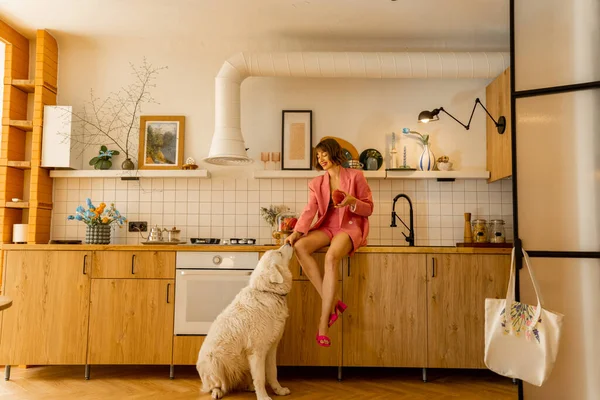 身穿粉色衣服的女人放松地坐在厨房的桌子上 一边吃一边喂着她那只硕大的白狗 有宠物在家里消闲时间的妇女 — 图库照片