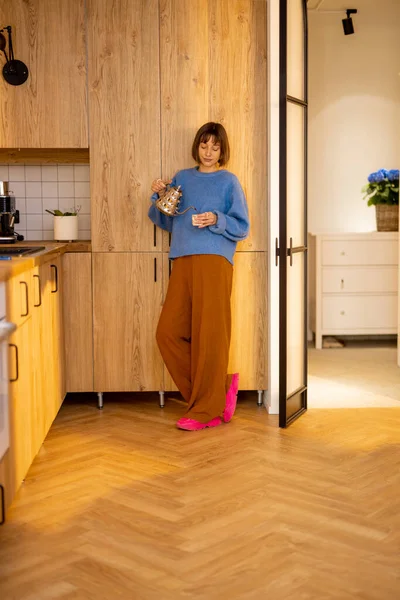 若い女性は 現代的なスタジオのアパートのキッチンにケトルとカップで立って余暇を過ごします 家庭生活とウェルネスの概念 — ストック写真
