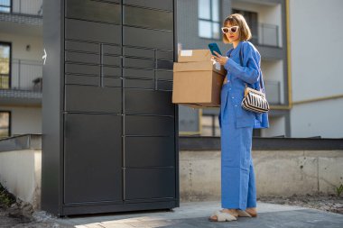 Genç bir kadın otomatik postane makinesinden paketleri alıp apartmanın yakınında duruyor. Otomatik dolaplara hızlı teslimat kavramı