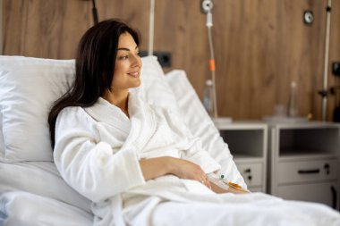 Kolunda kateter olan beyaz elbiseli güzel bir kadın hastane koğuşunda dinleniyor. Konforlu tıbbi süitte kadın hasta