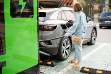Genç bir kadın açık hava şarj istasyonunda hızlı şarj olan bir araç için kablo takıyor. Elektrikli araba şarjı, elektrik ve yeşil enerji kavramı