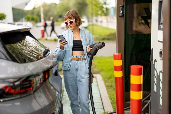年轻女子将在公共电站的车辆上为电动汽车充电 她将带插头的电缆用于快速充电和使用电话 城市快速充电器概念和新技术 — 图库照片