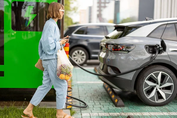 年轻妇女提着网袋中的一些杂货走到自己的电动车前 在车站外充电 可持续生活方式和绿色能源的概念 — 图库照片