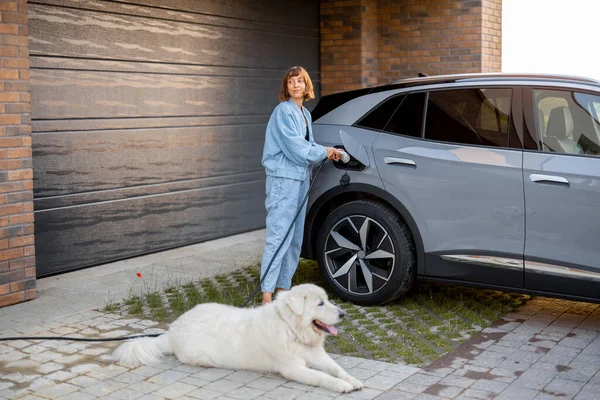 年轻女人和她可爱的狗站在她家车库附近时给电动车充电 现代快乐生活的概念 — 图库照片