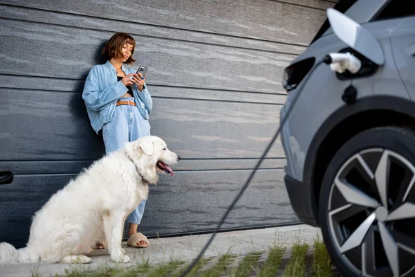 女人用手机靠在自家车库的门上 旁边有一只白色的狗 一辆电动车在前头充电 — 图库照片