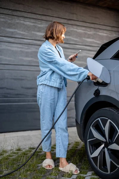 年轻女子站在她家车库门边拿着智能手机时 把枪插在车里充电 国产汽车收费的绿色及廉价能源概念 — 图库照片