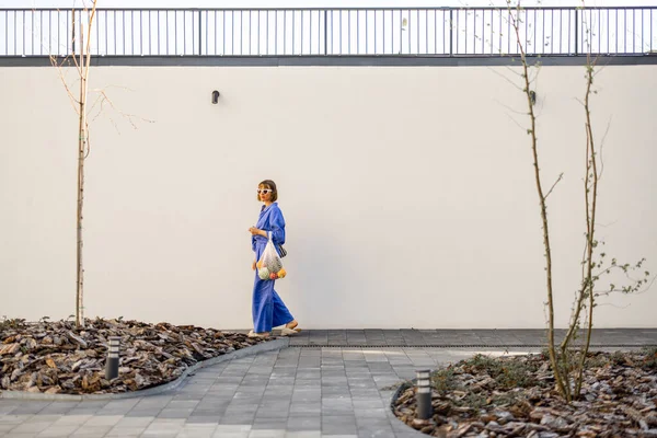 青いパジャマの女性は白い壁の背景の新鮮な果物でいっぱいのメッシュバッグで屋外を歩く 持続可能性と現代生活の概念 — ストック写真