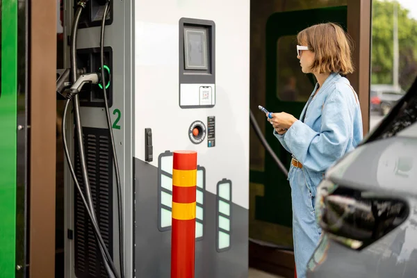 stock image Young woman going to charge electric car on vehicle public power station, scanning QR code with phone for paying. Concept of modern technologies for charging electric cars