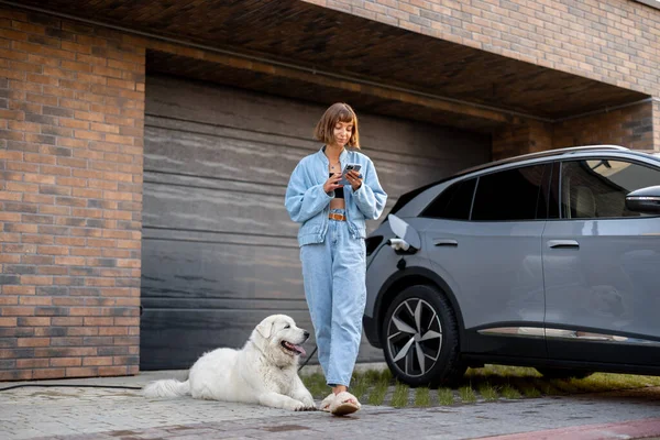 年轻女人和她的狗站在一起 用智能手机给她家车库附近的电动车充电 现代生活方式和可持续性的概念 — 图库照片