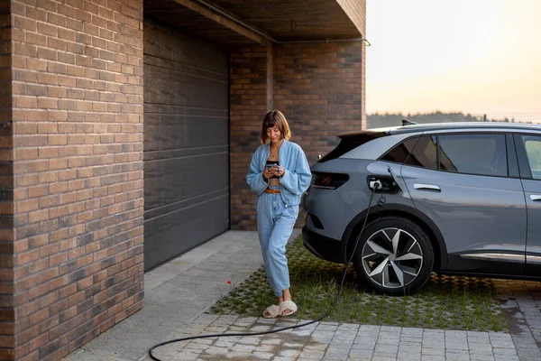 年轻妇女在自家车库附近给电动车充电时使用智能手机 现代生活方式和可持续性的概念 — 图库照片