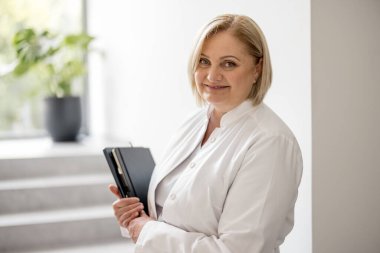 Klinikteki merdivenlerde, medikal önlüklü orta yaşlı hemşire portresi. Kapalı alanda klasörü olan güzel beyaz bir sağlık görevlisi.