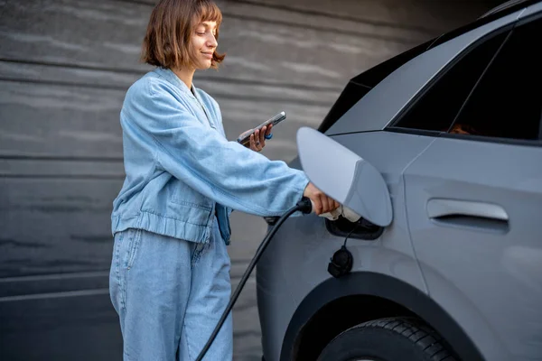 年轻女子站在她家车库门边拿着智能手机时 把枪插在车里充电 国产汽车收费的绿色及廉价能源概念 — 图库照片