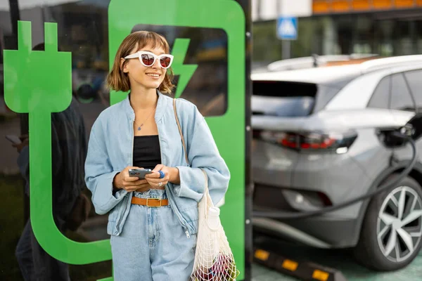 若い女性は いくつかの食品とスマートフォンやメッシュバッグで 彼女の電気自動車が屋外の公共駅で充電されるのを待って立っている 持続可能なライフスタイルとグリーンエネルギーの概念 — ストック写真