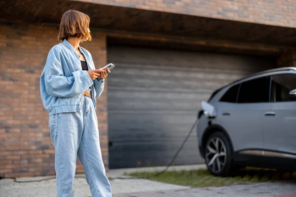 年轻妇女在给自家车库附近的电动车充电时 用的是智能手机 技术概念和电动车 — 图库照片