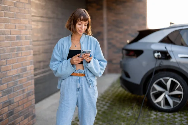 年轻妇女在自家车库附近给电动车充电时使用智能手机 现代生活方式和可持续性的概念 — 图库照片