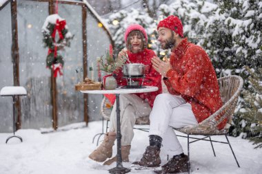 Erkek ve kadın fondüyle romantik bir akşam yemeği yerken, aynı zamanda güzel dekore edilmiş karlı arka bahçede masanın başında oturuyorlar. Genç bir aile kış tatilini dışarıda kutluyor.