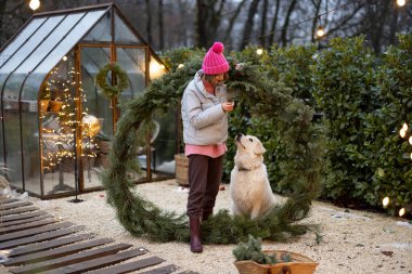 Genç kadın sevimli köpeğiyle oynuyor, arka bahçede büyük bir Noel çelengiyle dikilirken birlikte eğleniyor. Kış tatillerinde sevgi ve samimiyet kavramı