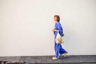 Mavi pijamalı bir kadın beyaz arka planda bir çanta dolusu taze meyveyle dışarıda yürüyor. Sürdürülebilirlik ve modern yaşam tarzı kavramı