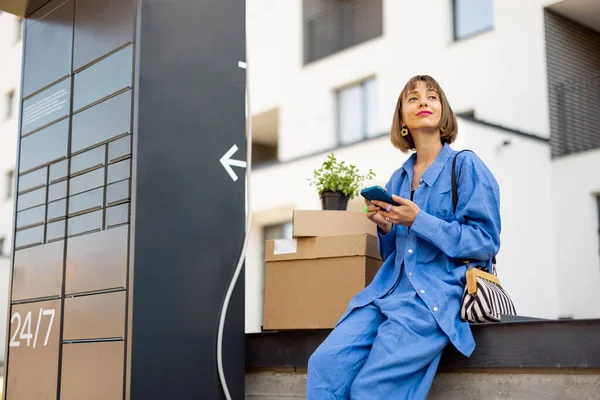 女性は郵便局の機械の近くに段ボールのパッケージと植木鉢と住宅地で携帯電話を使用して座っています 引渡し又は移転の概念 — ストック写真