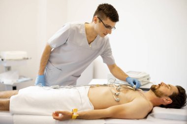 Genç kardiyolog, klinikteki orta yaşlı hastanın vücuduna yapışarak kardiyogram yapıyor.