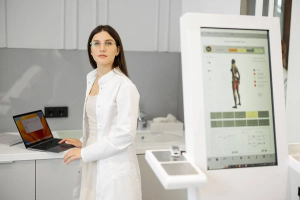 Young doctor works on laptop while standing near body analyzer machine at beauty salon. Concept of modern technologies in medicine and beauty industry