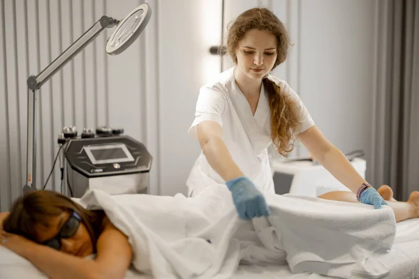 Young worker of a beauty salon covers woman with towel after laser epilation procedure