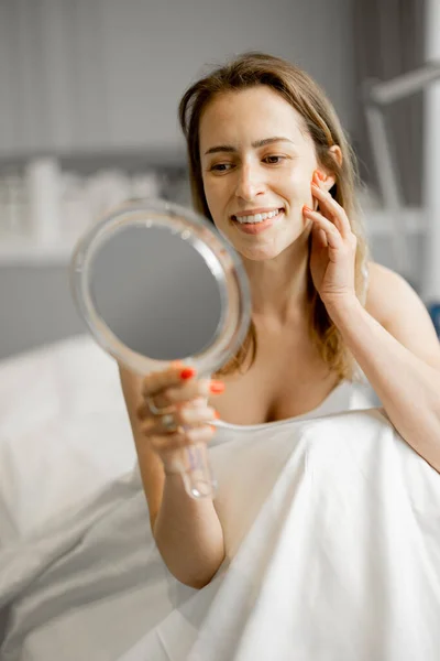 Happy woman looks in mirror on her face after beauty procedures while sitting at salon. Concept of beauty and facial care