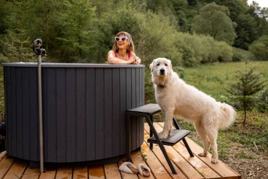 Bir kadın, sevimli köpeğiyle dağlardaki evinde dinlenirken dışarıda jakuzide banyo yapıyor. Eğlenme ve evcil hayvanlarla vakit geçirme kavramı.